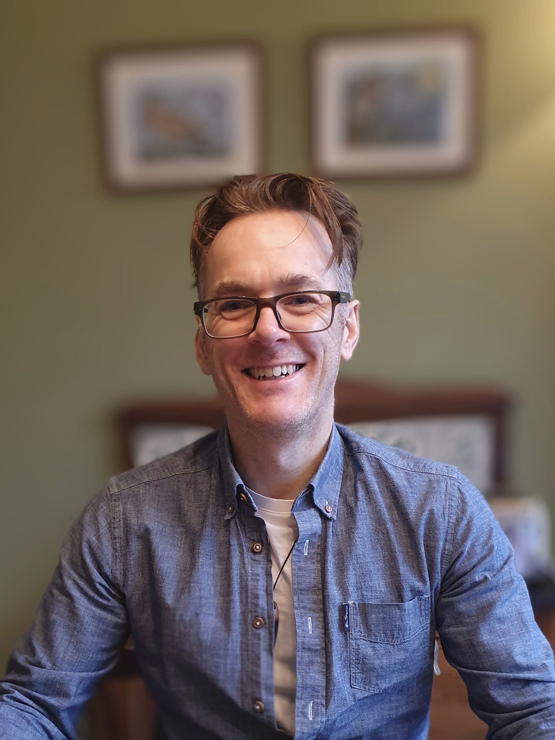 A smiling white man, facing the camera, black glasses, brown hair and a blue shirt.